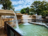 A multilevel water feature with cascading waterfalls flows into a large pool surrounded by black metal railings. The structure is made of light brown concrete and is set within a park-like environment with lush green trees and a partly cloudy blue sky.