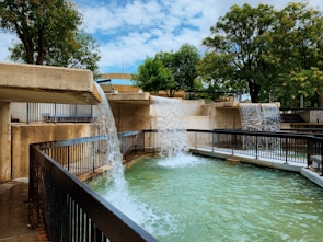 A multilevel water feature with cascading waterfalls flows into a large pool surrounded by black metal railings. The structure is made of light brown concrete and is set within a park-like environment with lush green trees and a partly cloudy blue sky.
