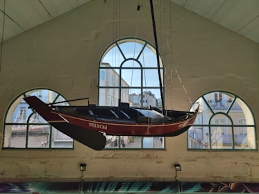 A wooden boat is suspended from the ceiling of a building with arched windows. The boat's name is visible on the side. Through the windows, a street view of colorful buildings can be seen.