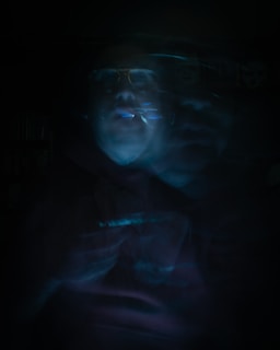 A blurred, eerie image featuring a human figure that appears ghostly with fragmented, overlapping features. The image is dark with low visibility, and the figure seems to be enveloped in a dim blue and greenish light.