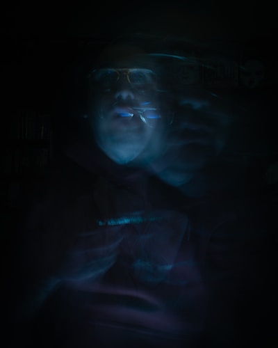 A blurred, eerie image featuring a human figure that appears ghostly with fragmented, overlapping features. The image is dark with low visibility, and the figure seems to be enveloped in a dim blue and greenish light.