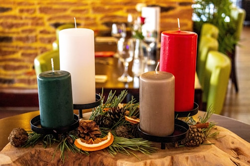 A set of four large pillar candles in red, white, green, and gray is placed on a wooden base. Pine cones, dried orange slices, and evergreen branches are used as decorative elements around the candles. The background shows a rustic interior with a yellow brick wall and green chairs.