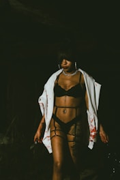 A person stands in dim lighting wearing black lingerie and a white garment splattered with red stains. They hold a knife in one hand, and their expression appears serious. The dark background adds to the mysterious atmosphere.