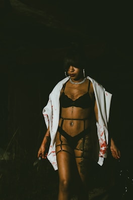 A person stands in dim lighting wearing black lingerie and a white garment splattered with red stains. They hold a knife in one hand, and their expression appears serious. The dark background adds to the mysterious atmosphere.
