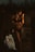 A person in dark lingerie poses on stone steps, partially illuminated by dim light. Nearby, a light blue shirt with a red design is draped over the steps, along with a single high-heeled shoe. The overall setting appears to be outdoors, with silhouettes of trees or foliage in the background.