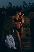 A woman in black lingerie stands on stone steps at dusk, with trees in the background. She is accessorized with large hoop earrings and striking red lipstick. Nearby, on the steps, are a pair of high-heeled shoes and a white garment featuring a red pattern.