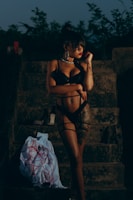 A woman in black lingerie stands on stone steps at dusk, with trees in the background. She is accessorized with large hoop earrings and striking red lipstick. Nearby, on the steps, are a pair of high-heeled shoes and a white garment featuring a red pattern.