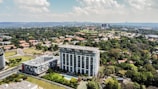 Aerial view of a cityscape featuring a large, modern building surrounded by greenery and smaller residential structures. In the background, the skyline extends with more buildings amidst a lush landscape. The sky is partly cloudy, providing a bright and vibrant atmosphere.