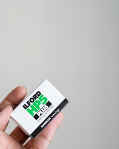 A close-up of a hand holding a box of Ilford HP5 Plus black and white film against a plain light-colored background. The box is primarily white with bold black and green text showcasing the brand and type of film.