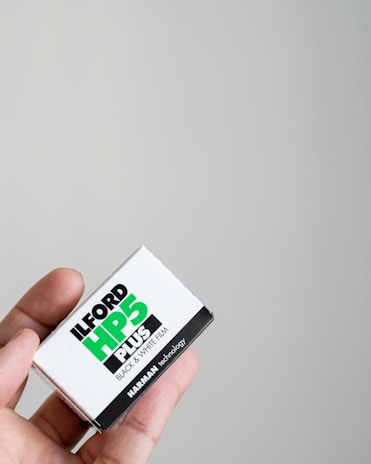 A close-up of a hand holding a box of Ilford HP5 Plus black and white film against a plain light-colored background. The box is primarily white with bold black and green text showcasing the brand and type of film.