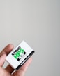 A close-up of a hand holding a box of Ilford HP5 Plus black and white film against a plain light-colored background. The box is primarily white with bold black and green text showcasing the brand and type of film.