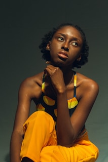 A person with short curly hair sits pensively, resting their chin on their hand. They wear a colorful top with yellow, blue, and orange patterns and bright yellow pants. The background is dark and minimalistic, emphasizing the subject's thoughtful expression.