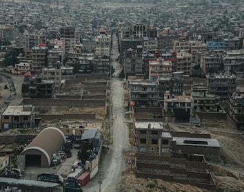 A dense urban area with numerous multi-story buildings lined along a central road. The architecture is varied, with some unfinished structures and colorful rooftops, surrounded by rough terrain and sparse vegetation. The environment appears arid and dusty, typical of a developing city.