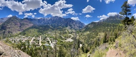 A panoramic view of a mountainous landscape with a small town nestled in a valley. The foreground shows a hiking trail with scattered trees and bushes. The midground features a cluster of buildings, grid-like streets, and clear roads. The background consists of towering, rugged mountains covered in greenery, partially capped with snow, set against a bright blue sky with fluffy white clouds.