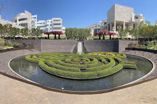 A beautifully landscaped garden features an intricate circular hedge maze surrounded by a reflective water feature. The setting is complemented by modern architectural buildings in the background, characterized by their light stone facades and large windows. Vibrant pink-flowered trees accent the greenery and add visual interest to the scene.