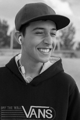 A young person is smiling broadly while wearing a dark hoodie with the 'Vans' logo and a cap. They have earphones plugged in, and the background is blurred, suggesting an outdoor setting.