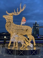 An illuminated sculpture of a stag and a fox made from a framework of glowing lights stands prominently. The stag has elaborate antlers, and a bird is perched on its back. The backdrop features a bridge with Gothic-style towers, including visible lights hinting at evening or night. The fence surrounds the sculpture, and a person is observing the installation.