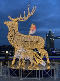 An illuminated sculpture of a stag and a fox made from a framework of glowing lights stands prominently. The stag has elaborate antlers, and a bird is perched on its back. The backdrop features a bridge with Gothic-style towers, including visible lights hinting at evening or night. The fence surrounds the sculpture, and a person is observing the installation.