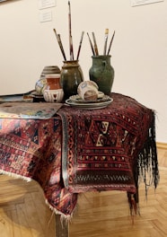A table is covered with a richly patterned red and black textile, on which sits a collection of artist's materials. There are several ceramic jars and mugs containing paintbrushes, each varying in size and color. Additionally, there is a palette with mixed paint colors and a few decorative pottery items, including a small vase and a bowl.