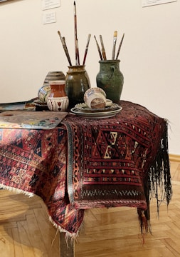 A table is covered with a richly patterned red and black textile, on which sits a collection of artist's materials. There are several ceramic jars and mugs containing paintbrushes, each varying in size and color. Additionally, there is a palette with mixed paint colors and a few decorative pottery items, including a small vase and a bowl.