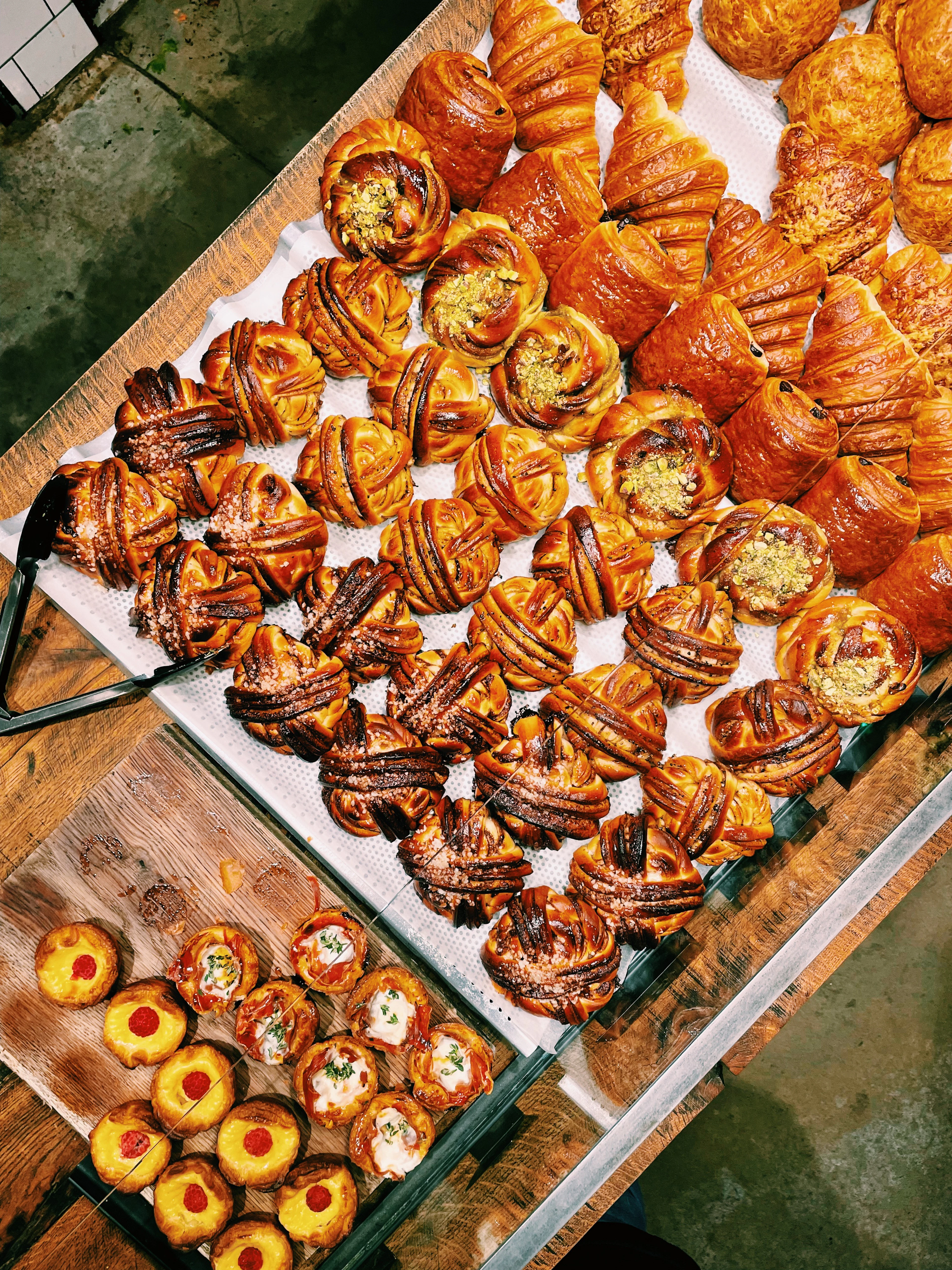 Trays of Estonian bakery pastries seen from above: golden croissants, twisted cinnamon kringel-style buns and small jam- and cheese-topped tarts laid out on a bakery counter