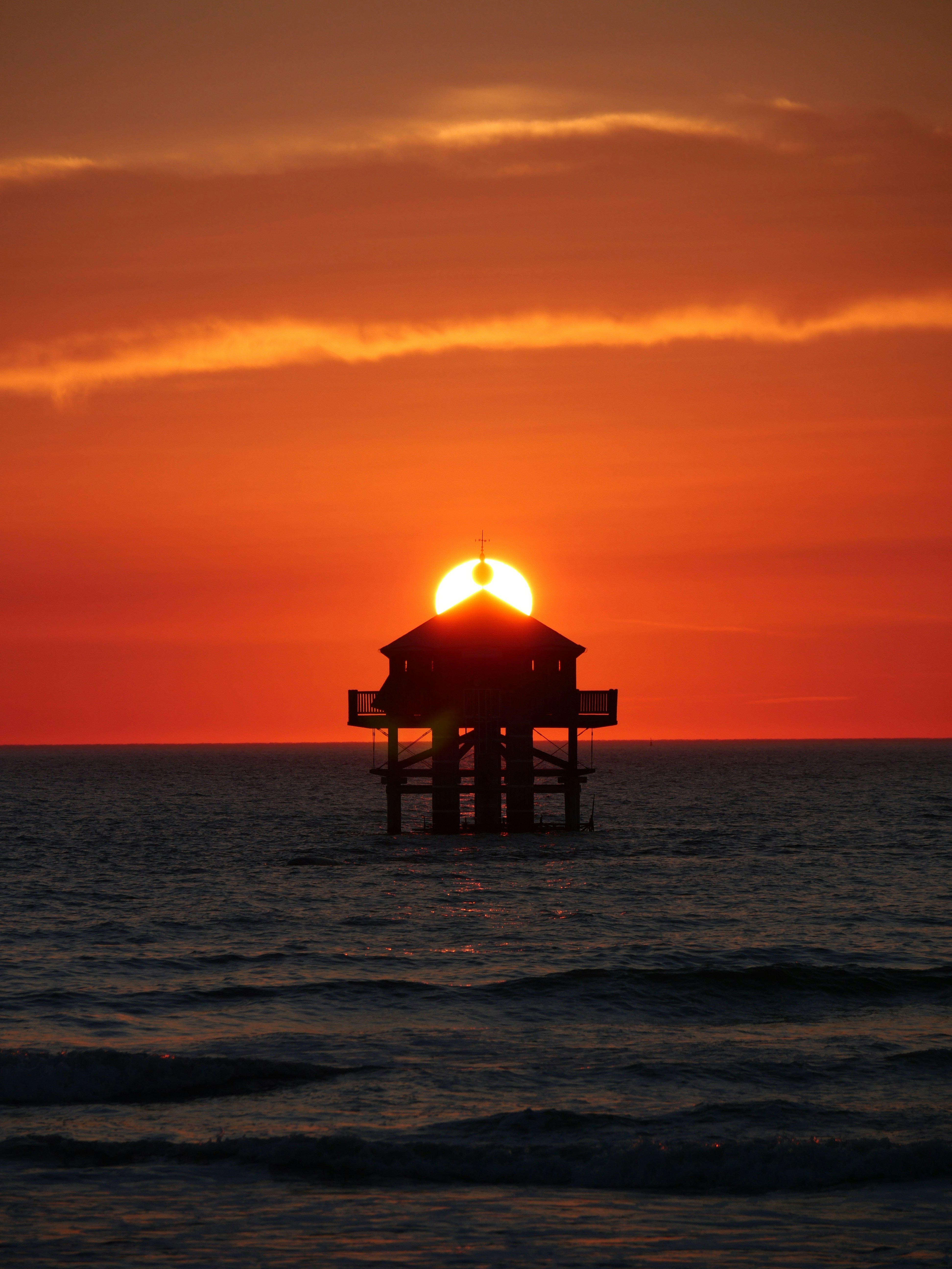 Silhouette of a pier on stilts rising from calm water against a fiery sunset, with the sun aligned behind the pier's roof to form a halo. Photograph.