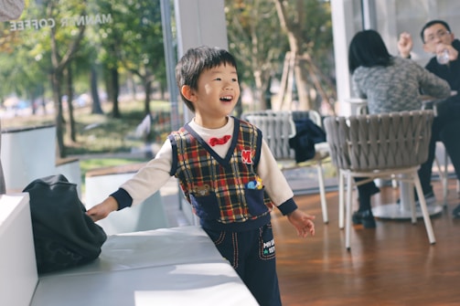 A young child with a cheerful expression stands inside a cafe, wearing a plaid vest over a light sweater. Sunlight filters through large windows illuminating the space, and in the background, two adults sit at a table, engaged in conversation.