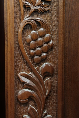 Intricate wooden carving featuring a grapevine design. The artwork includes a cluster of grapes and swirling leaves, showcasing detailed craftsmanship with a textured background. The natural wood grain adds to its aesthetic appeal.