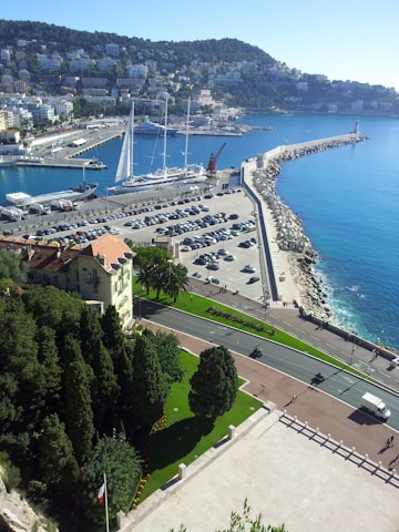 A coastal cityscape featuring a marina with several large sailboats docked. The water is a vibrant blue, and the coastline is lined with a breakwater extending into the sea. The hillside is densely populated with buildings surrounded by lush greenery. In the foreground, there's a well-maintained garden and a road with moving vehicles.