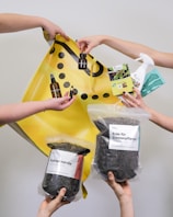 Multiple hands are holding various gardening and plant care products such as soil bags, plant nutrients, a spray bottle, and small bottles with liquids against a plain background with a yellow shopping bag.