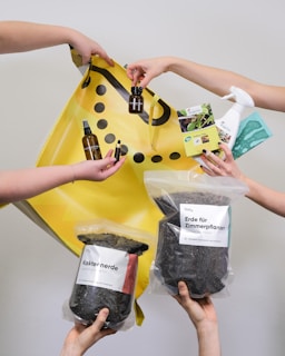 Multiple hands are holding various gardening and plant care products such as soil bags, plant nutrients, a spray bottle, and small bottles with liquids against a plain background with a yellow shopping bag.