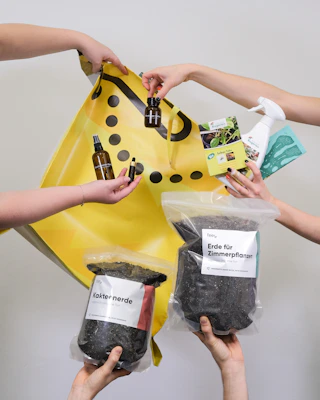 Multiple hands are holding various gardening and plant care products such as soil bags, plant nutrients, a spray bottle, and small bottles with liquids against a plain background with a yellow shopping bag.