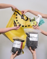 Multiple hands are holding various gardening and plant care products such as soil bags, plant nutrients, a spray bottle, and small bottles with liquids against a plain background with a yellow shopping bag.