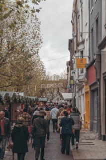 A bustling street market with numerous people walking down a narrow street lined with stalls and trees. The atmosphere is lively, and the stalls are adorned with garlands and decorations, suggesting a festive or holiday theme. Buildings on the right side display various signs, while the overcast sky adds to the cozy, autumnal feel.