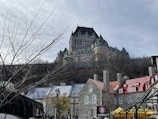 A grand, castle-like hotel with turrets and spires sits atop a hill, overlooking a collection of historic stone buildings with colorful roofs. Bare tree branches fill the foreground, hinting at a wintry setting.