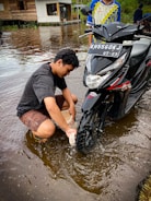 A person is washing a motorcycle tire while sitting in shallow water. The motorcycle has a visible license plate, and another person stands beside it. In the background, there's a wooden house on stilts next to a body of water, which reflects the surroundings.