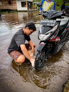 A person is washing a motorcycle tire while sitting in shallow water. The motorcycle has a visible license plate, and another person stands beside it. In the background, there's a wooden house on stilts next to a body of water, which reflects the surroundings.