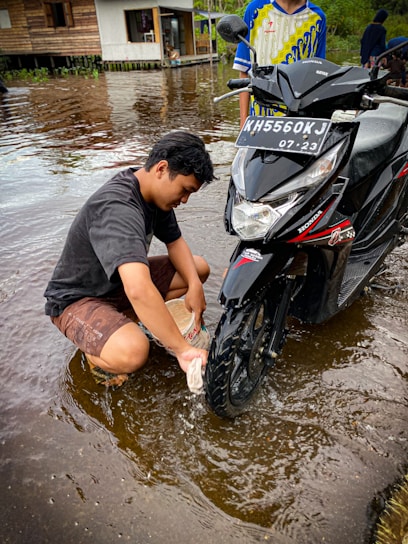 A person is washing a motorcycle tire while sitting in shallow water. The motorcycle has a visible license plate, and another person stands beside it. In the background, there's a wooden house on stilts next to a body of water, which reflects the surroundings.