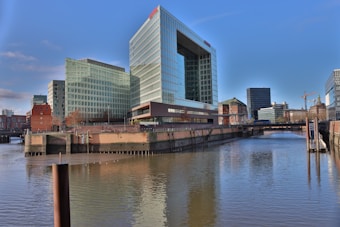 A modern glass office building with multiple stories is situated on the edge of a calm body of water. The reflection of the structure is visible in the water. In the background, there are additional buildings, a crane, and clear blue sky. The area seems to be part of an urban setting with elements of both modern and traditional architecture.