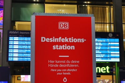 A bright red sign with the text 'Desinfektionsstation' prominently displayed, indicating a place to sanitize hands. Below is a smaller text in German and English explaining that hands can be disinfected here. The background features digital departure boards with various train information, suggesting a train station setting. There's also a glimpse of a storefront with illuminated signage.