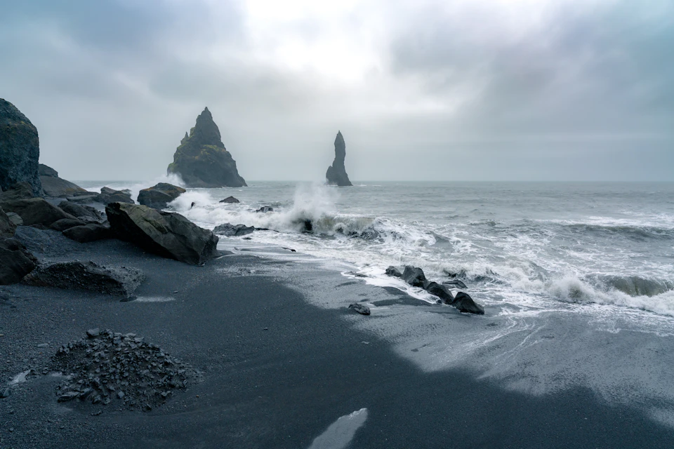 The image depicts a rocky beach with two large, dark gray rock formations standing out against the light blue sky and choppy ocean waves. The rocks are positioned on either side of the frame, creating a sense of balance within the composition. The water is a darker shade of blue than usual, suggesting that it may be choppy or rough due to the windy conditions.
