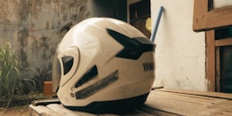 A white motorcycle helmet with black accents is resting on a wooden surface. Visible is the brand name Yamaha and the model name XMAX. The background includes a weathered concrete wall, some greenery, and a partially visible wooden window frame.