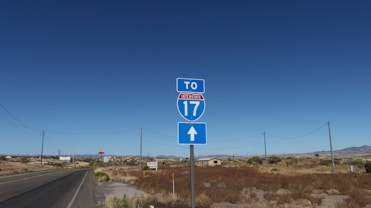 A road sign indicating the direction to Interstate 17 stands by the side of an empty road. The landscape is arid with dry vegetation, utility poles, and a distant view of structures and mountains under a clear blue sky.