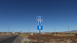 A road sign indicating the direction to Interstate 17 stands by the side of an empty road. The landscape is arid with dry vegetation, utility poles, and a distant view of structures and mountains under a clear blue sky.