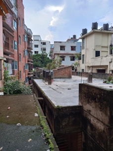 A view of an urban residential area with multiple mid-rise apartment buildings. The buildings have a variety of architectural styles, with some featuring red brick exteriors and others with white or cream facades. Rooftops are visible, equipped with water tanks and greenery. Vegetation and small trees are seen along the edges, and the sky is partly cloudy.
