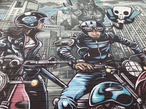 A vibrant mural depicts two motorcyclists wearing helmets and leather gear, riding through a stylized cityscape. The background features tall buildings and a large robot with a skull and crossbones symbol. The mural uses a comic book style with bold outlines and dynamic poses.