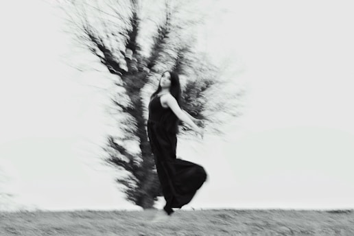 A blurred image of a person dancing or moving gracefully outside, dressed in a flowing black dress against a backdrop of a leafless tree. The movement creates a sense of dynamism and fluidity.
