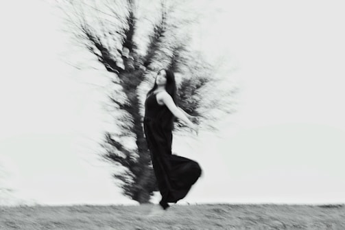 A blurred image of a person dancing or moving gracefully outside, dressed in a flowing black dress against a backdrop of a leafless tree. The movement creates a sense of dynamism and fluidity.