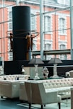 A modern interior space featuring elegant seating arrangements with tufted armchairs and sofas. A table with decorative lamps and vases is prominently placed. In the background, a large window overlooks an exterior with a brick building facade. A contemporary sculpture of human figures holding circular rings is displayed against a dark cylindrical structure.