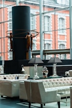 A modern interior space featuring elegant seating arrangements with tufted armchairs and sofas. A table with decorative lamps and vases is prominently placed. In the background, a large window overlooks an exterior with a brick building facade. A contemporary sculpture of human figures holding circular rings is displayed against a dark cylindrical structure.